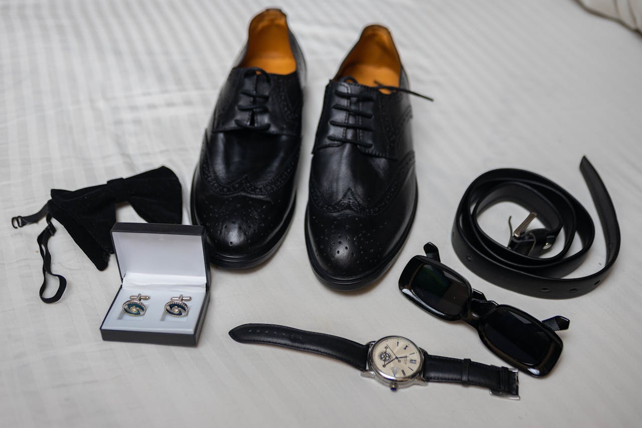 Flat lay of stylish men's accessories including shoes, sunglasses, and cufflinks on a white background.